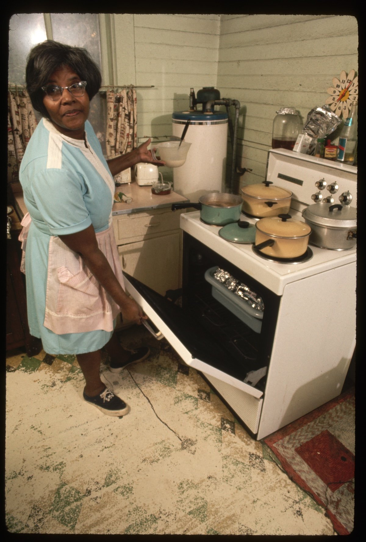 An older woman in a blue dress and apron is standing in a vintage kitchen, opening the oven door while holding a bowl. Several pots are on the stovetop, and the kitchen has a retro, homey atmosphere.