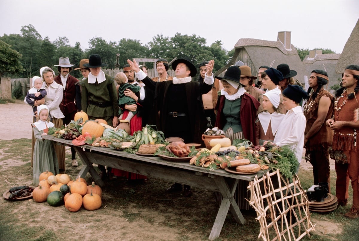 People in historical Pilgrim and Native American costumes gather around a large outdoor table filled with autumn harvest food, as a man in black raises his arms, appearing to give thanks. Thatched buildings and trees are in the background.