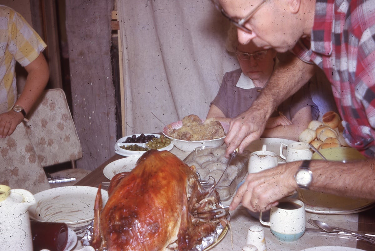 A person carves a roasted turkey at a dinner table set with various dishes, capturing the warm, nostalgic feel often seen in vintage Thanksgiving photos. Two other people are seated at the table, one partially visible and one wearing glasses and a hat.