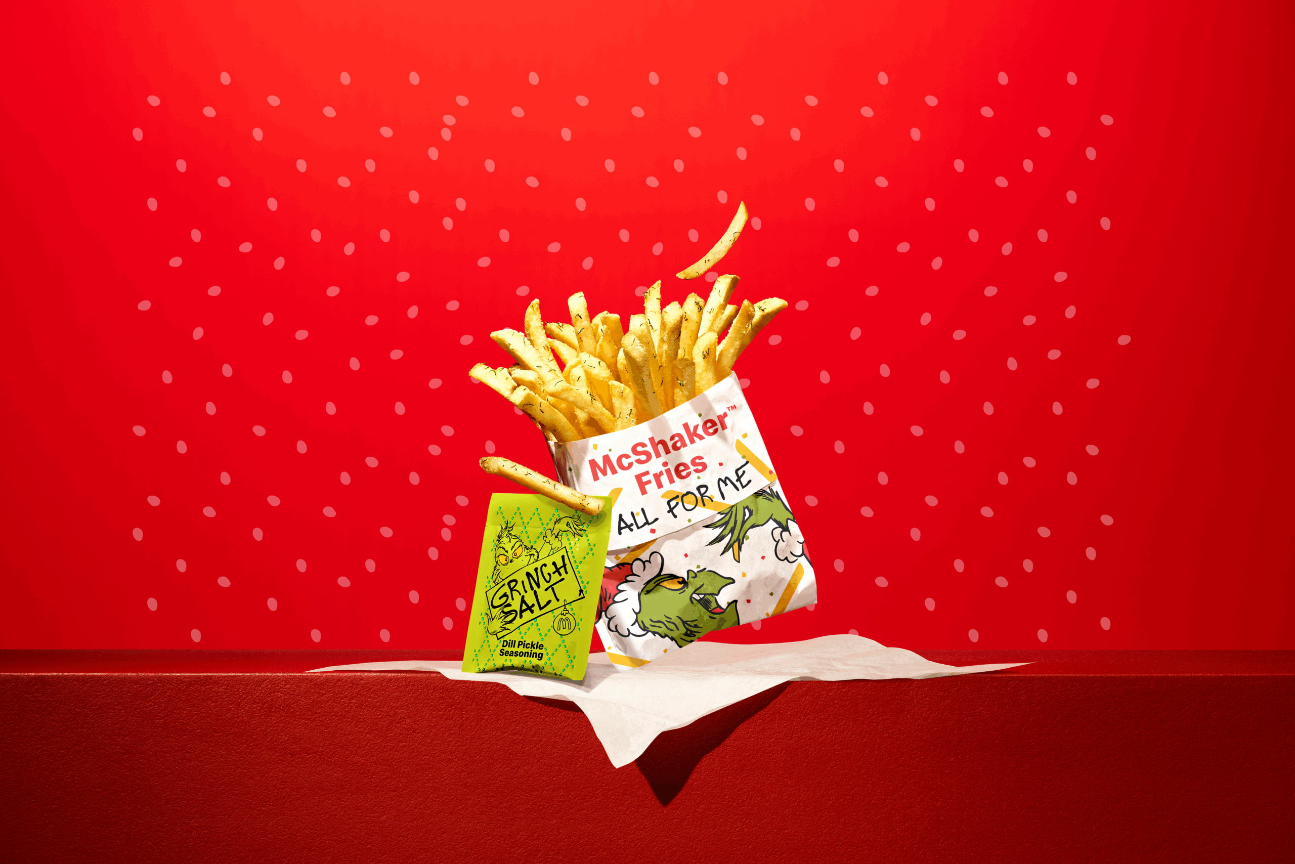 A McShaker Fries package with cartoon illustrations sits on a napkin, with fries spilling out and a small green packet labeled “Crunchy Salt” against a bright red background with white dot accents.