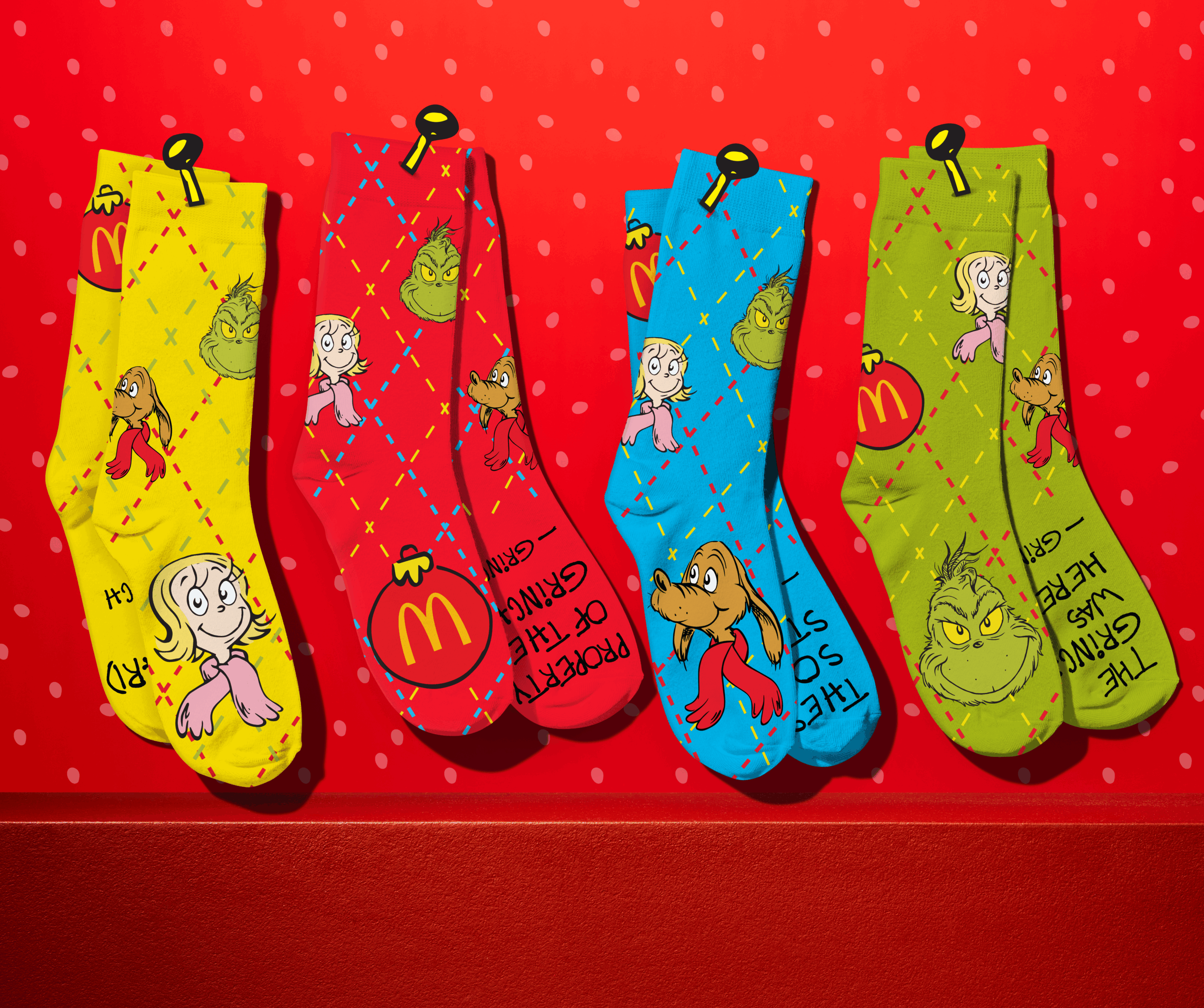 Four colorful socks with Dr. Seuss' Grinch-themed characters and McDonald's logos hang against a red, polka-dotted background. The socks are yellow, red, blue, and green.