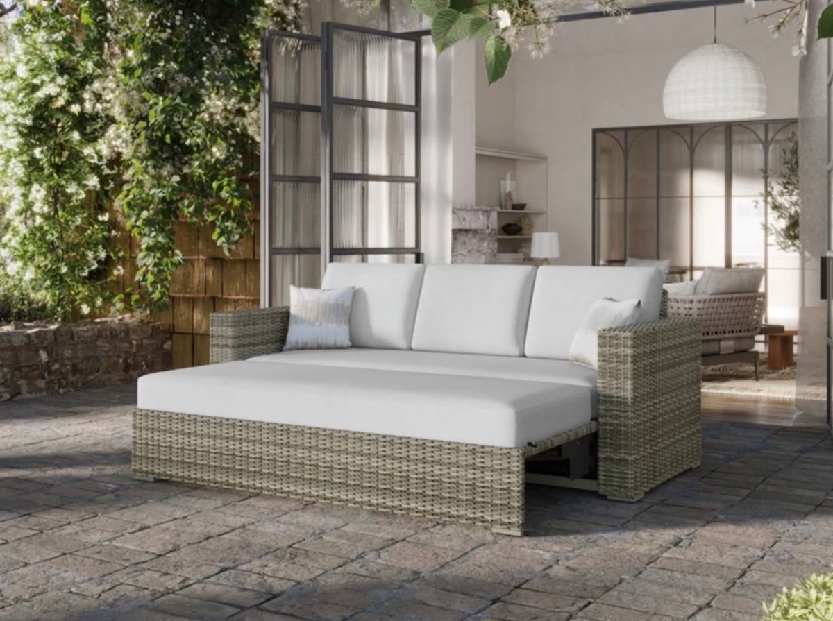 A modern outdoor patio with a wicker sectional sofa featuring white cushions, set on a stone floor. The area is surrounded by greenery and flowers, with glass doors open to a cozy, light-filled indoor space.