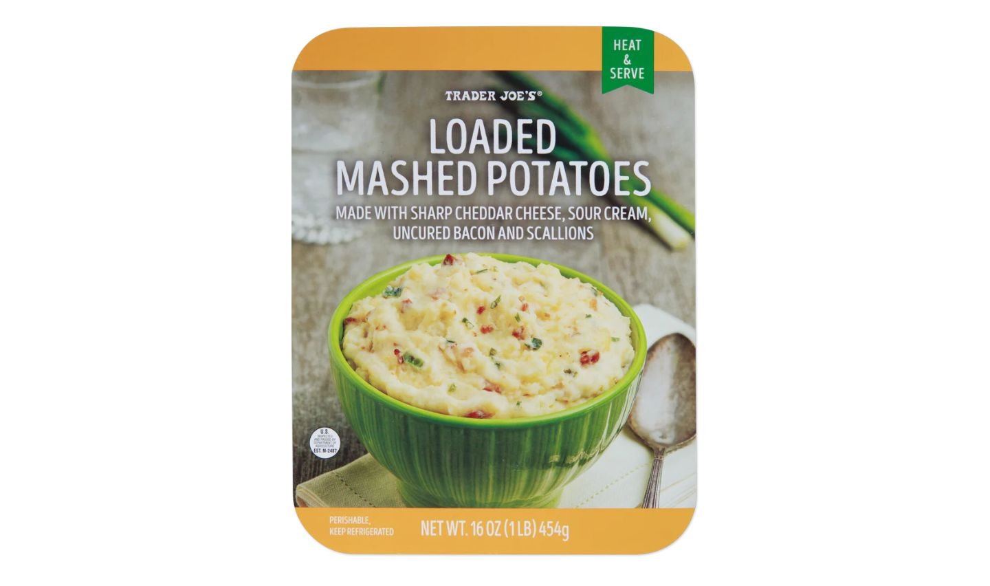 Packaging of Trader Joe’s Loaded Mashed Potatoes, showing a green bowl filled with mashed potatoes mixed with cheese, bacon, and scallions. The text highlights sharp cheddar, sour cream, bacon, and scallions.