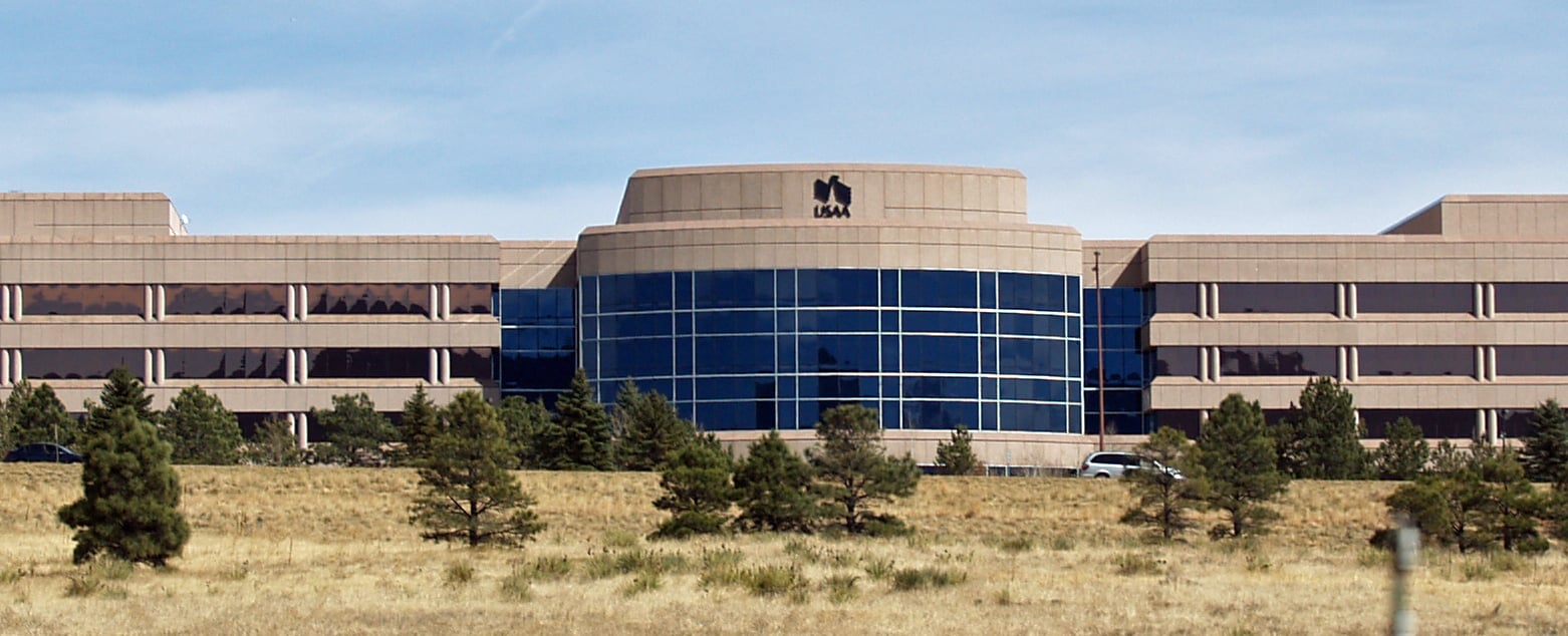 A large, modern office building with blue-tinted windows, tan walls, and a USAA logo on top, surrounded by trees and grass under a clear sky.