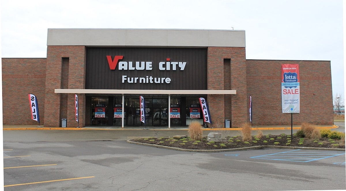 A Value City Furniture store with a brick exterior and large sign. Several "SALE" banners are displayed near the entrance, and the parking lot in front includes a marked accessible parking space.