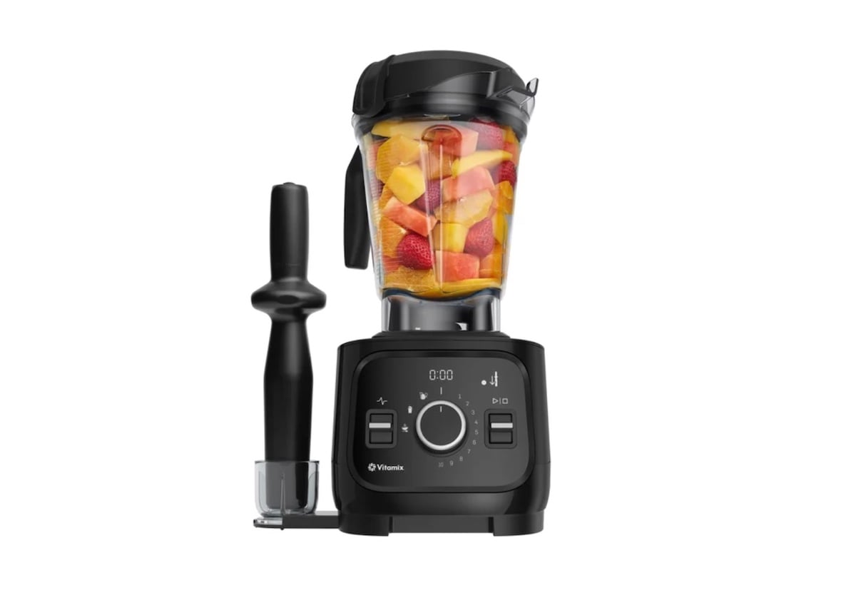 A black countertop blender filled with colorful chopped fruits, including strawberries, mango, and pineapple. A tamper tool is attached to the side, and the blender base features digital controls and a display screen.