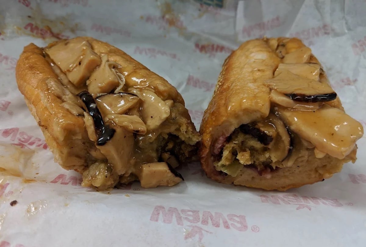 A sandwich cut in half, filled with thick slices of tofu, sauce, and what appears to be mushrooms, on a piece of wax paper with faint red text in the background.