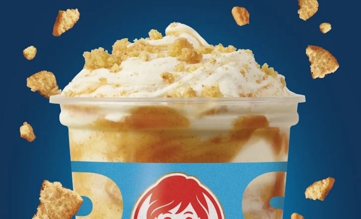 A Wendy's dessert cup filled with creamy ice cream swirled with golden caramel and topped with crumbled cookie pieces, set against a blue background with cookie crumbs scattered around.