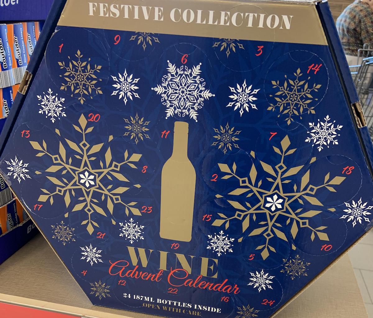 A festive hexagonal wine advent calendar box with gold and white snowflakes, numbered doors from 1 to 24, and the words “Festive Collection” and “Wine Advent Calendar” on a blue background.