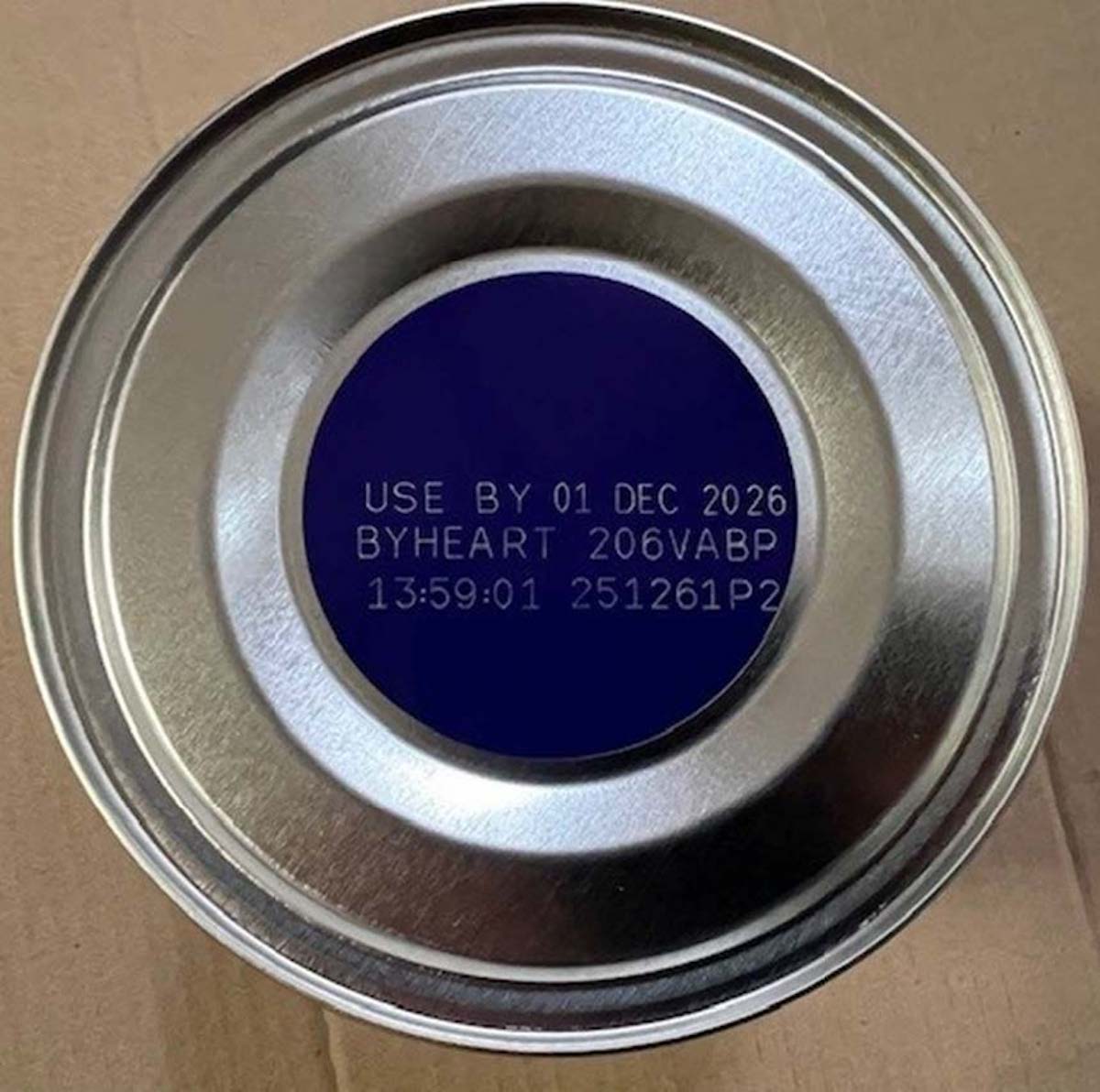 A close-up of the bottom of a metal can with text printed on it: "USE BY 01 DEC 2026 BYHEART 206VABP 13:59:01 251261P2." The can is placed on a cardboard surface.
