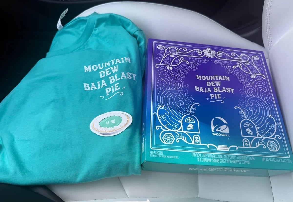 A turquoise T-shirt with "Mountain Dew Baja Blast Pie" text and a pie graphic sits next to a purple pie box with the same text and Taco Bell logo on a white car seat.