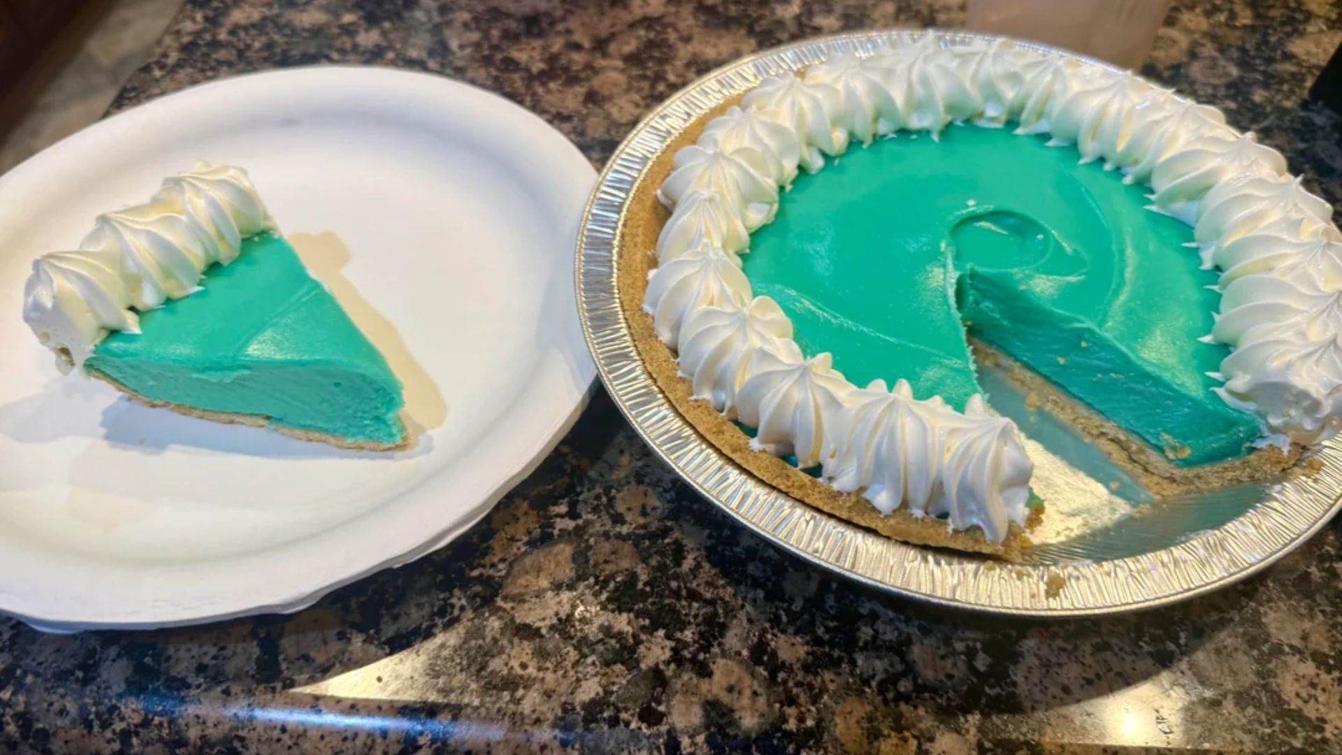 A whole blue pie with a graham cracker crust and whipped cream border sits on a counter; one slice has been served on a white paper plate next to it.