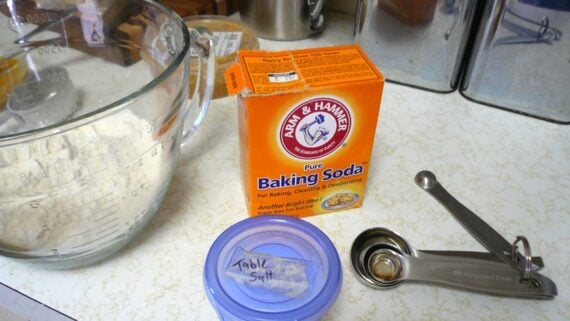 A box of Arm & Hammer baking soda sits on a kitchen counter next to a glass mixing bowl with flour, a container labeled “table salt,” and a set of metal measuring spoons.