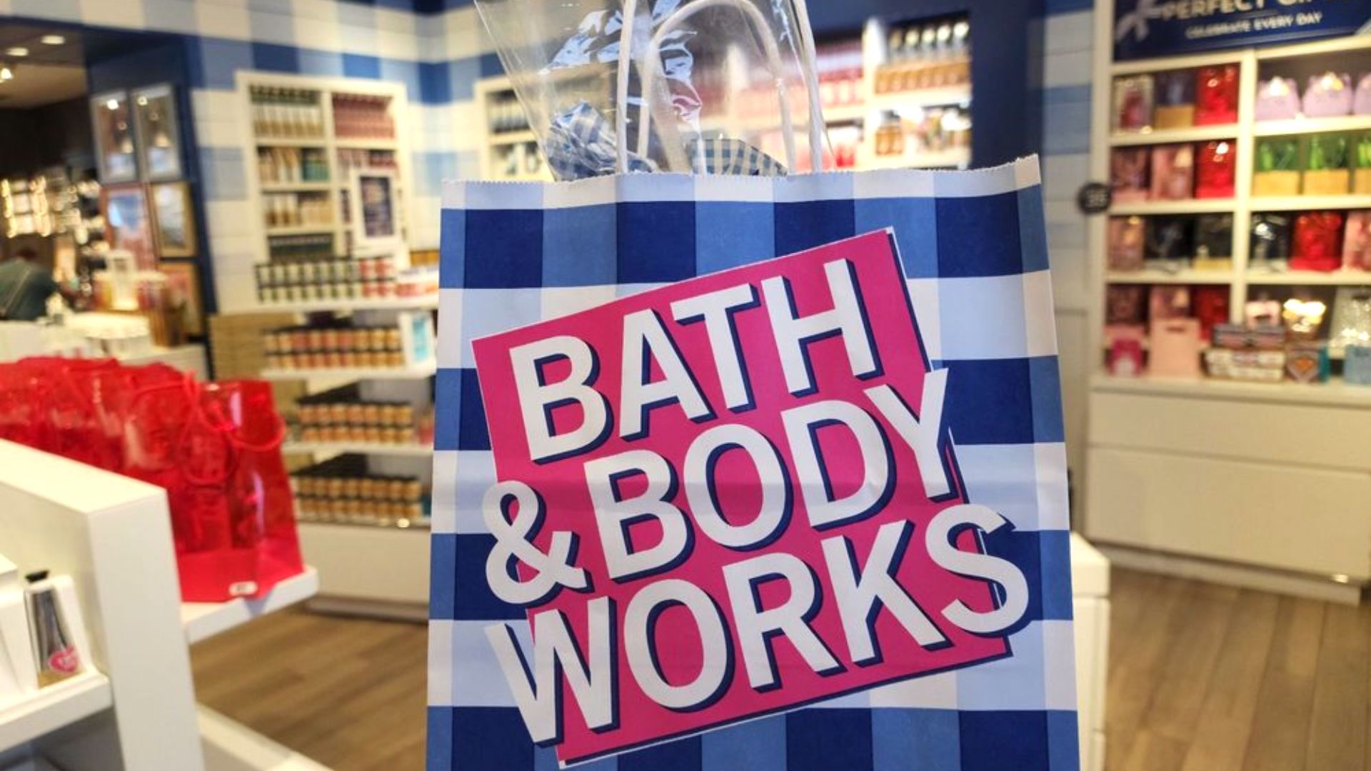 A close-up of a blue and white checkered Bath & Body Works shopping bag held inside a brightly lit store, with shelves displaying colorful products in the background.