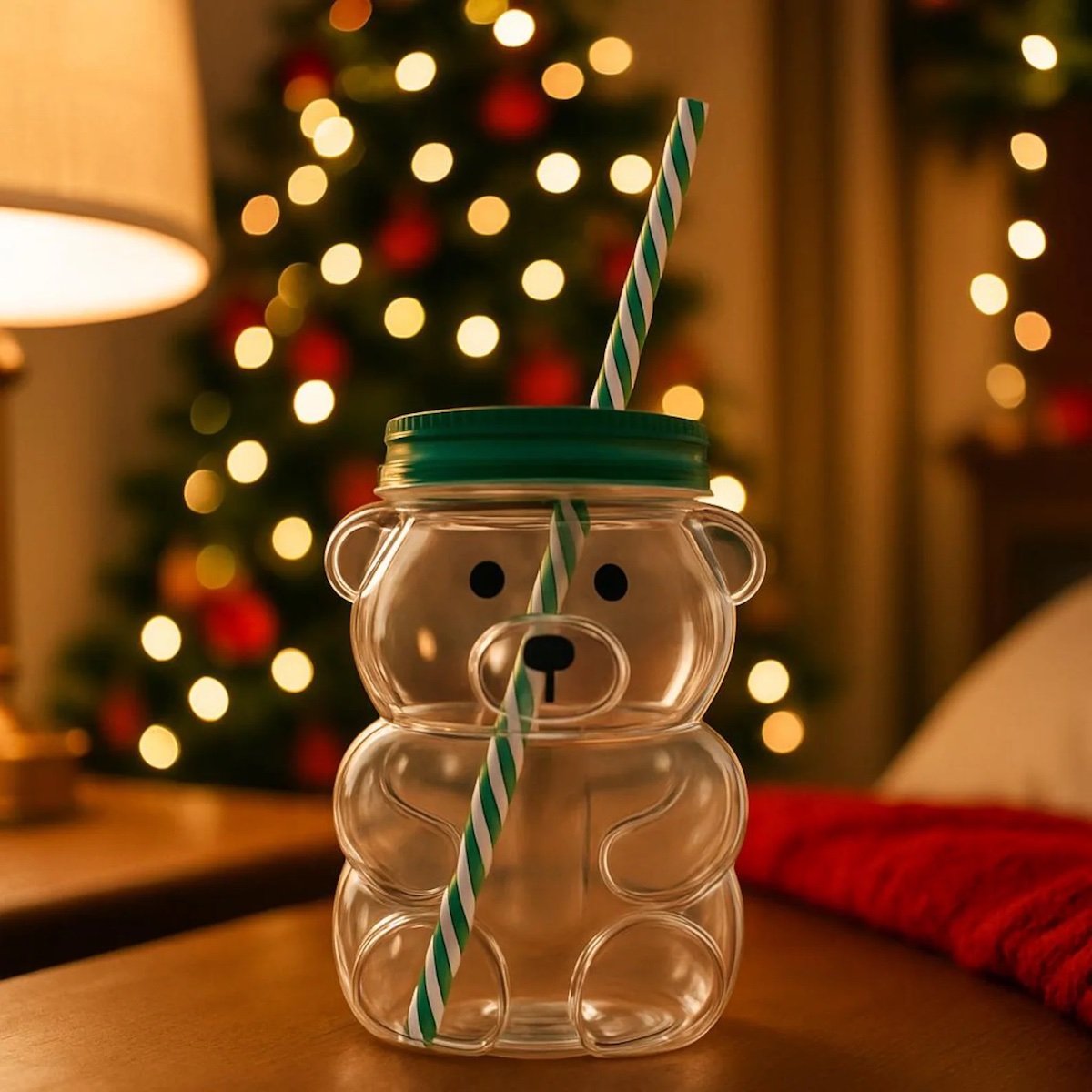 A clear bear-shaped cup with a green lid and striped straw sits on a table. In the background, a lit Christmas tree and festive decorations create a cozy holiday atmosphere.