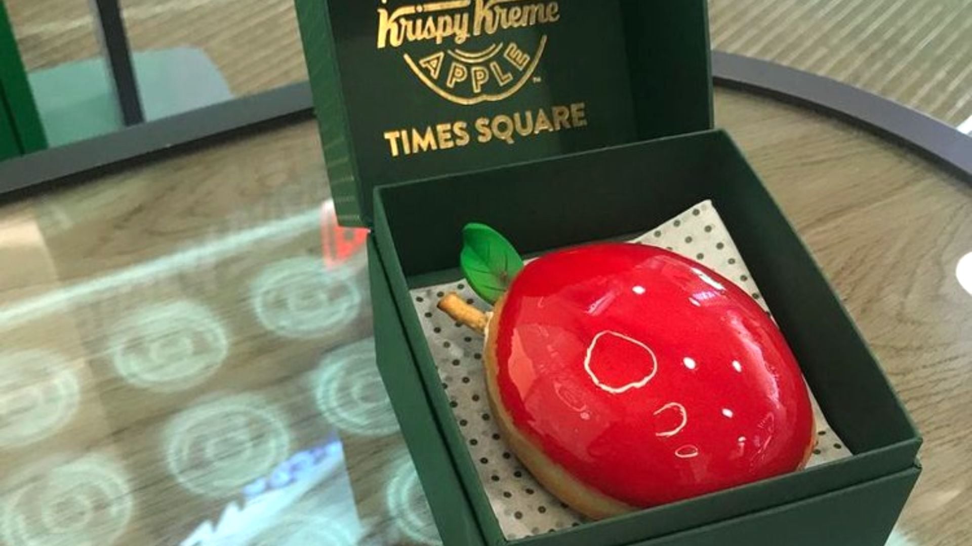 A shiny, red, apple-shaped donut with a green leaf and brown stem in a green Krispy Kreme Times Square box, resting on a dotted paper liner on a glass table.