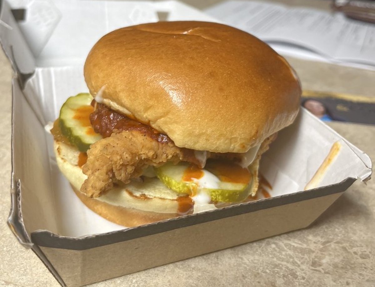 A crispy chicken sandwich with pickles, sauce, and a soft bun is served in an open cardboard takeout box on a countertop.