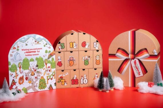 Three decorative holiday gift boxes are displayed on a red background, including an advent calendar with numbered doors, a colorful illustrated box with festive wishes, and a striped box with a bow, surrounded by mini trees and ornaments.