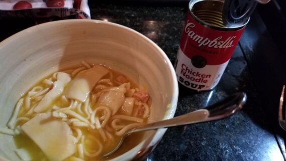 A bowl of chicken noodle soup with a spoon sits next to an open can of Campbell’s Chicken Noodle Soup on a dark countertop. Pieces of noodle, chicken, and possibly potatoes are visible in the soup.