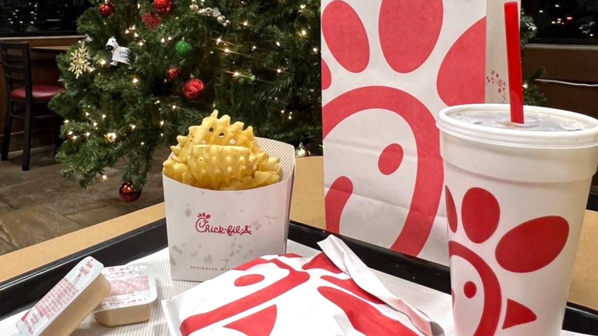 A tray with Chick-fil-A food items, including waffle fries, a wrapped sandwich, sauce containers, and a large drink cup, sits on a table. A decorated Christmas tree is visible in the background.