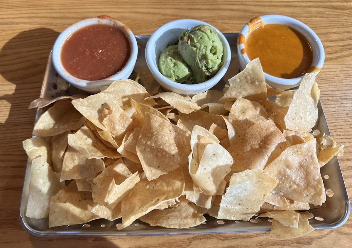A tray of crispy tortilla chips served with three dips: red salsa, guacamole, and a yellow cheese sauce, all in small bowls on a wooden table.