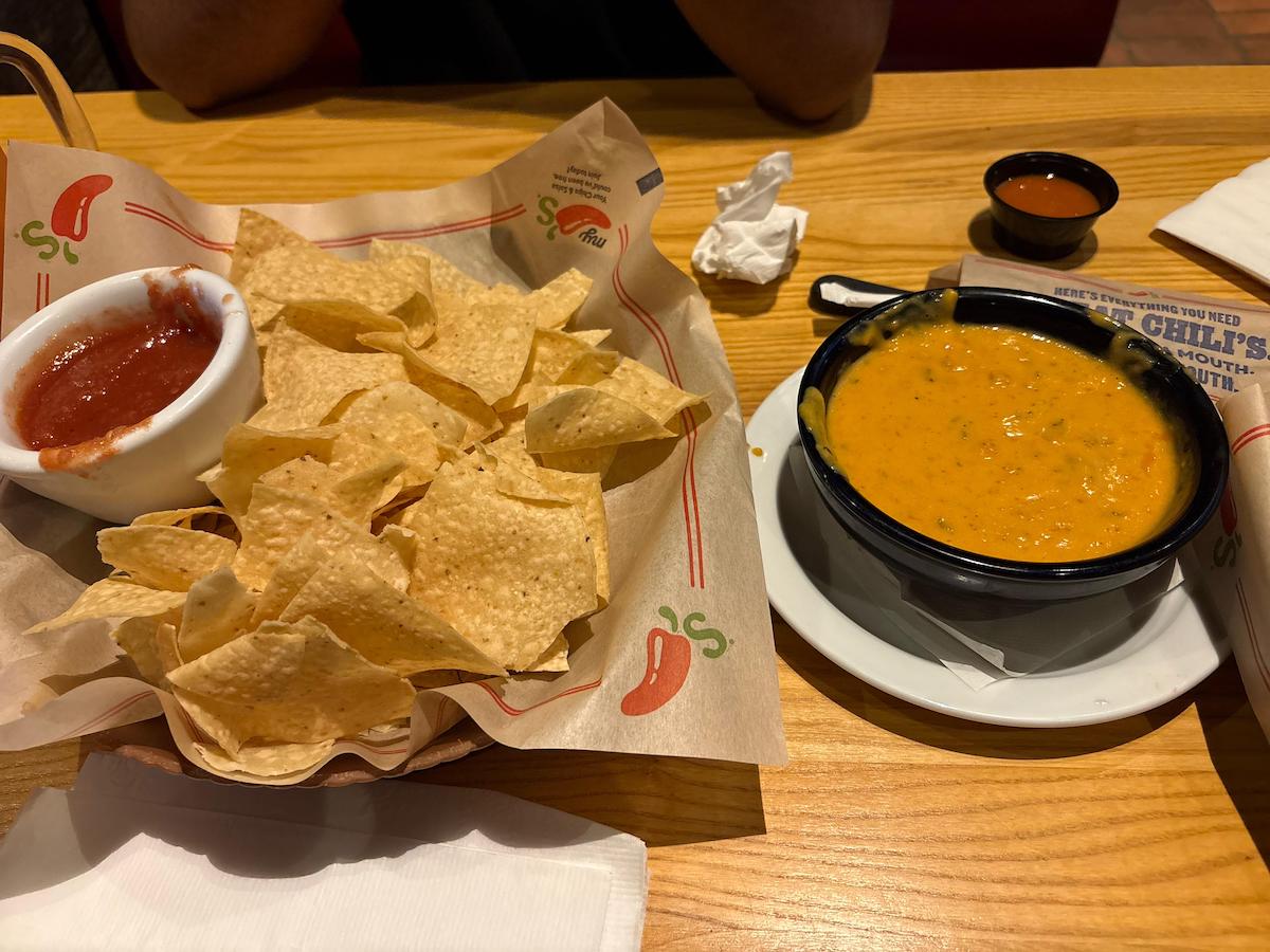 A basket of tortilla chips with a bowl of salsa sits next to a bowl of melted yellow queso dip on a wooden table at a restaurant. A napkin and small cup of sauce are also visible.