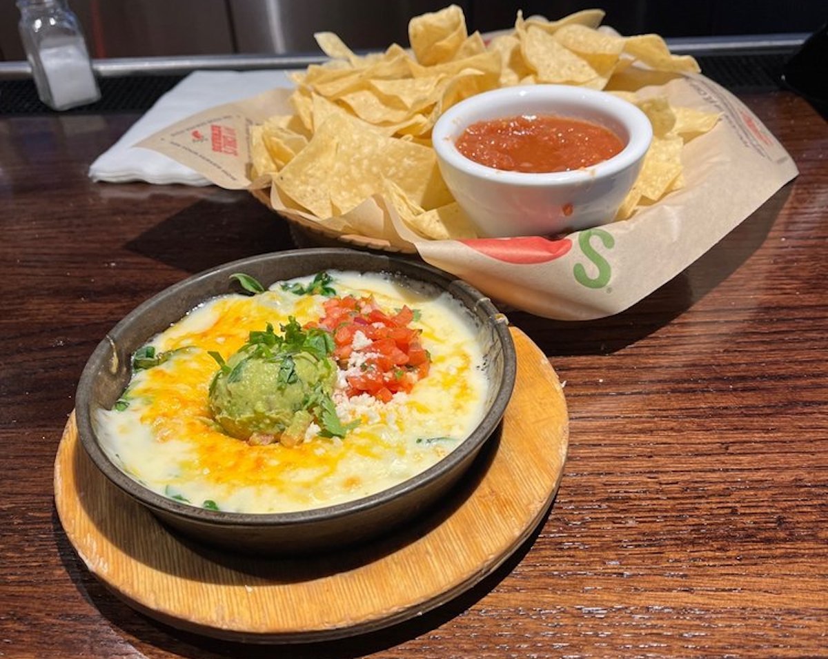 A bowl of cheesy dip topped with guacamole, tomatoes, and herbs sits on a wooden plate in front of a tray of tortilla chips and a small bowl of salsa on a wooden table.