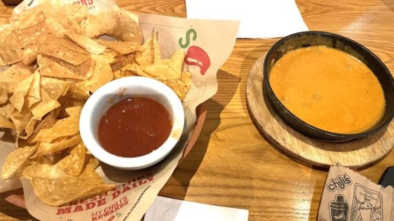 A basket of tortilla chips with a bowl of salsa sits next to a bowl of yellow cheese queso dip on a wooden table at a Chili's restaurant.