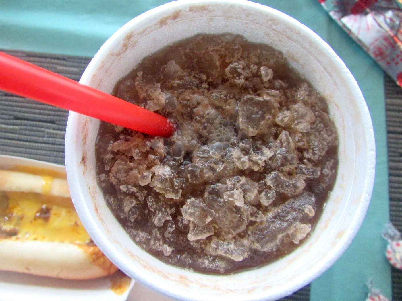 A close-up of a styrofoam cup filled with crushed ice and a dark soda, with a red straw inserted. Part of a hot dog with melted cheese is visible in the lower left corner.