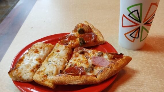A red plate with three slices of pizza and a breadstick sits on a beige table next to a white cup with a colorful geometric design.
