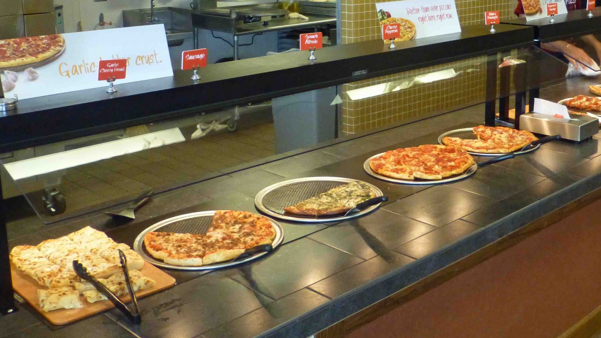 A buffet counter displays several varieties of pizza and breadsticks on trays, with serving tongs nearby. The pizzas are under a glass sneeze guard, and the kitchen area is visible in the background.