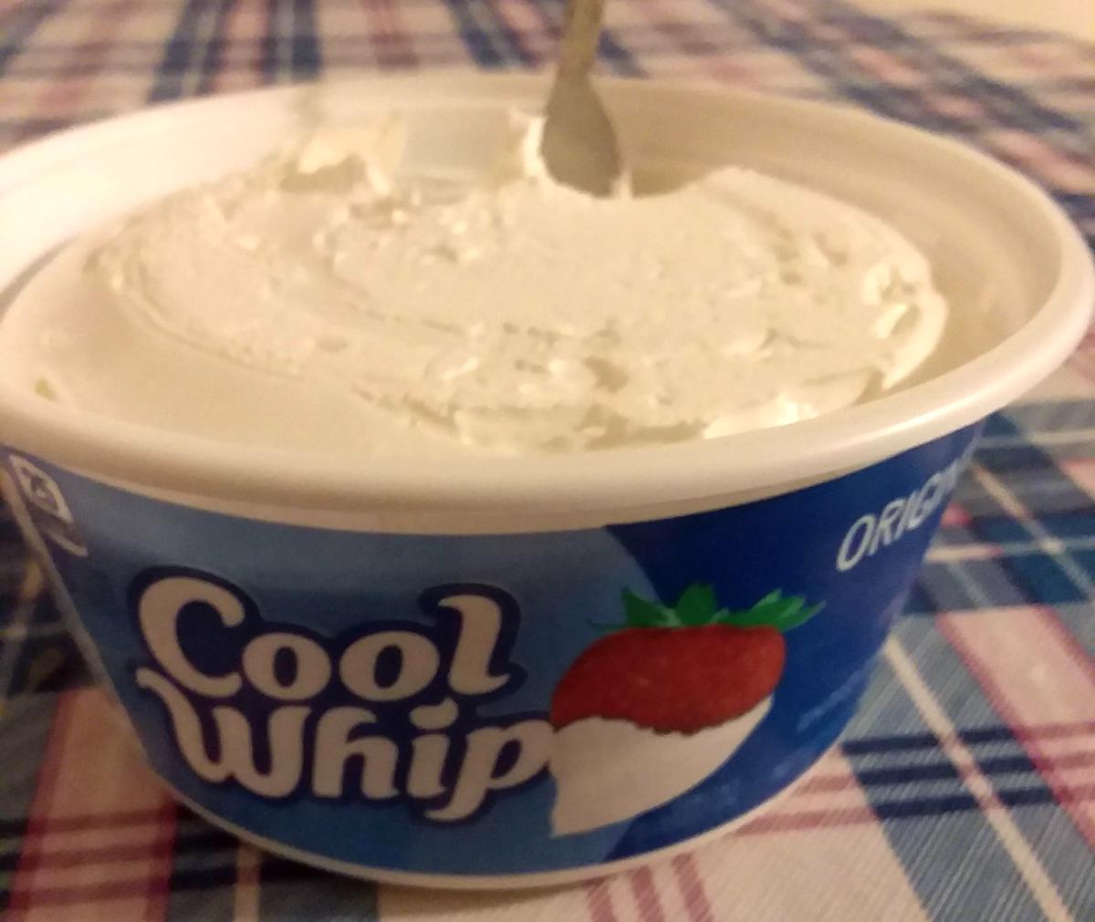 A tub of Cool Whip whipped topping with a spoon inside, placed on a plaid tablecloth. The blue container features the Cool Whip logo and an image of a strawberry with whipped topping.