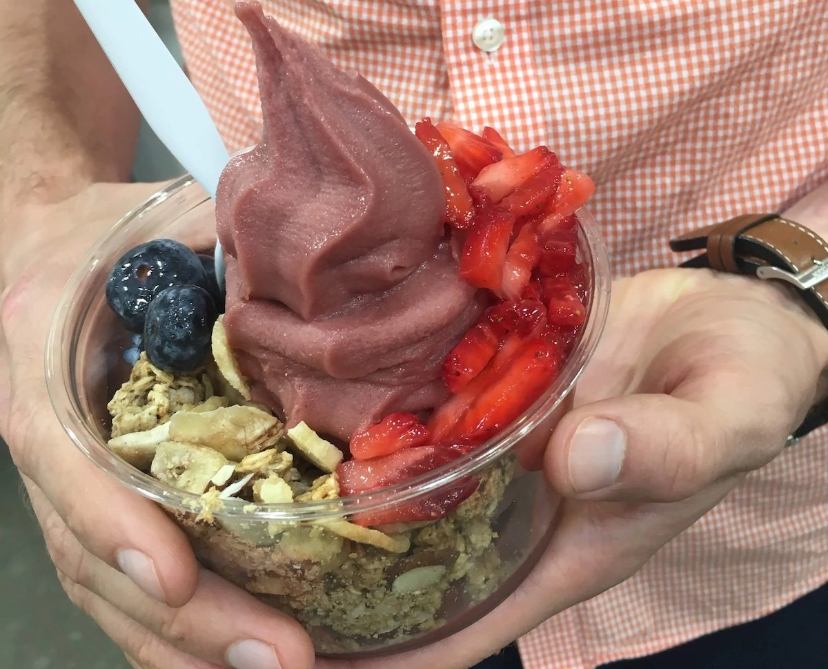 A person holding a cup of acai soft serve topped with granola, fresh blueberries, and sliced strawberries, with a spoon in the cup. The person is wearing an orange checkered shirt and a brown watch.