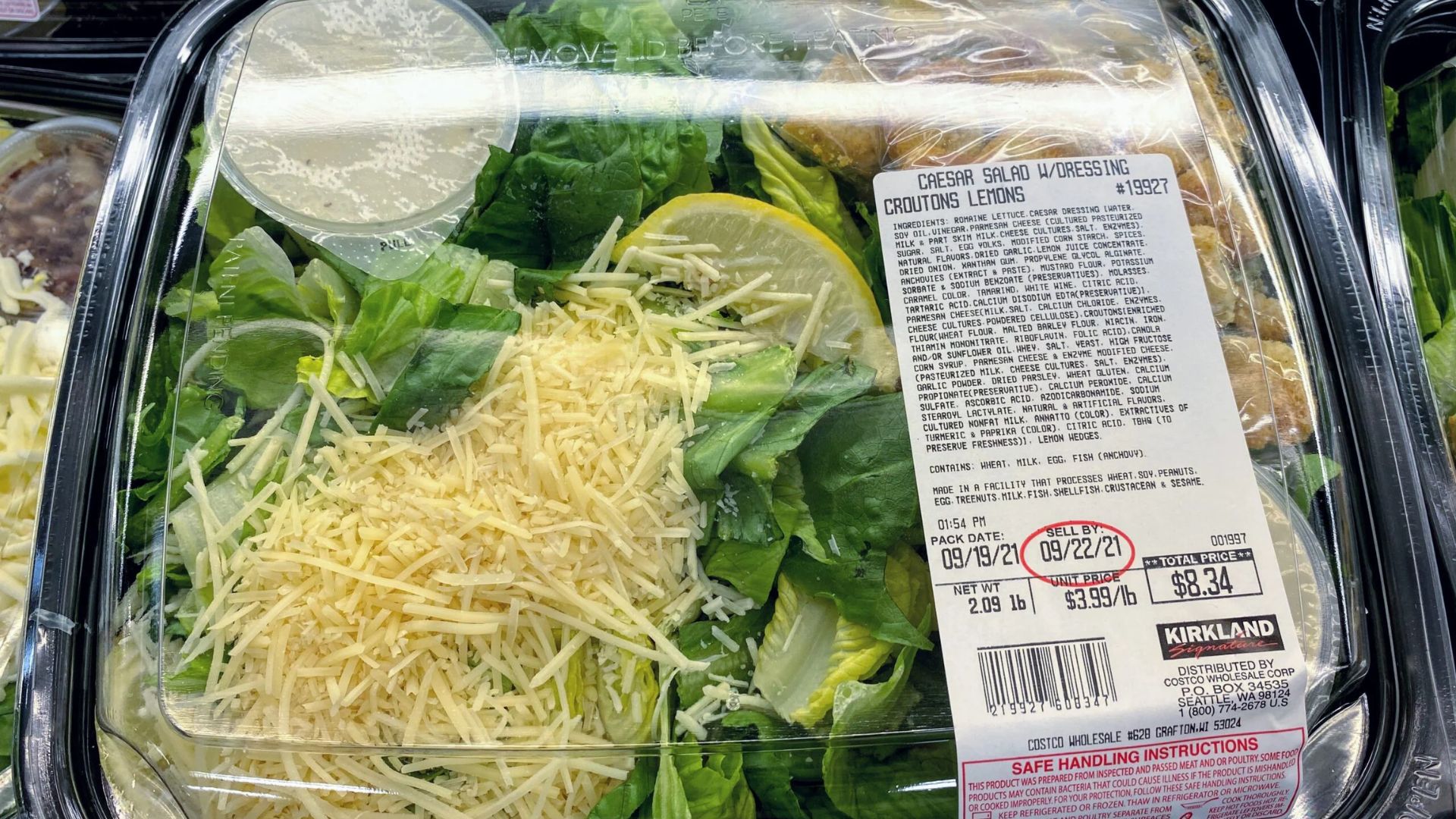 A plastic container of Kirkland Caesar salad with lettuce, shredded cheese, lemon slices, croutons, and dressing packet, labeled with nutrition and price information.