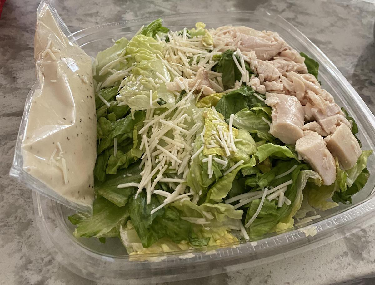 A plastic container holds chopped romaine lettuce, shredded Parmesan cheese, diced cooked chicken, and a packet of creamy salad dressing, all ready to be mixed for a fresh salad.