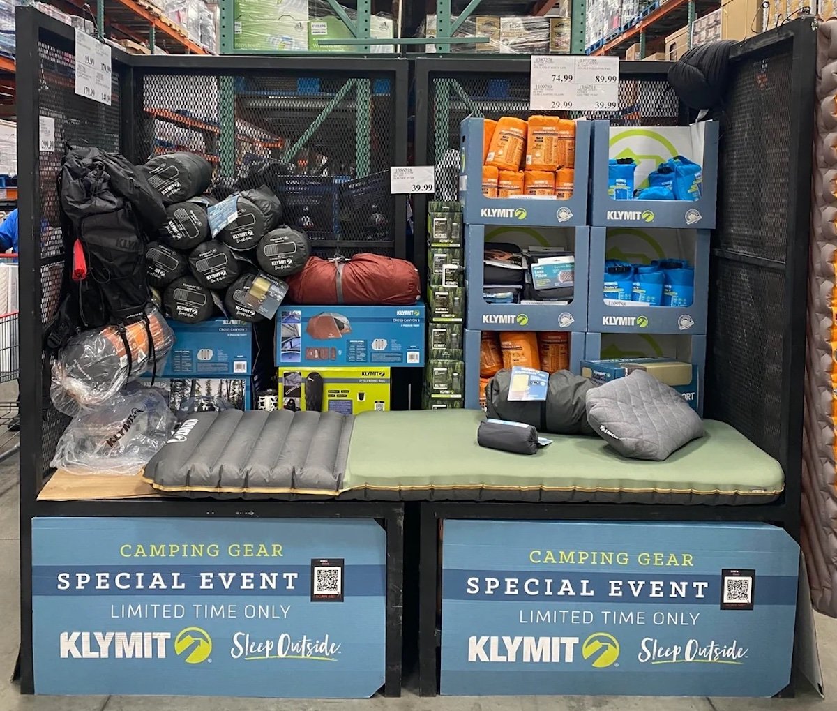 A store display features Klymit camping gear, including sleeping pads, pillows, sleeping bags, and tents. Signs read “Camping Gear Special Event - Limited Time Only.” The items are organized on benches and shelves.