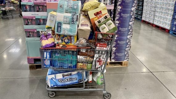 A shopping cart overflowing with bulk items such as toilet paper, paper towels, snacks, cleaning supplies, and household goods in a warehouse store aisle. Stacks of large packaged goods are visible in the background.