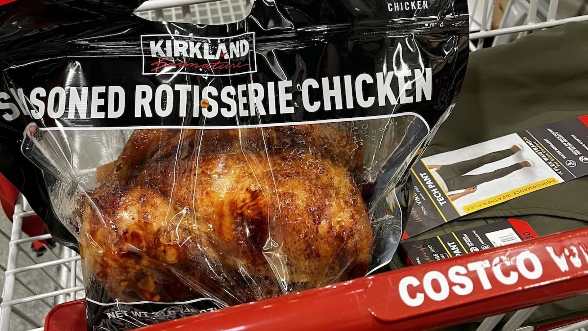A Kirkland Signature seasoned rotisserie chicken in a plastic bag sits in a Costco shopping cart along with packaged items. The red handle of the cart displays the Costco logo.