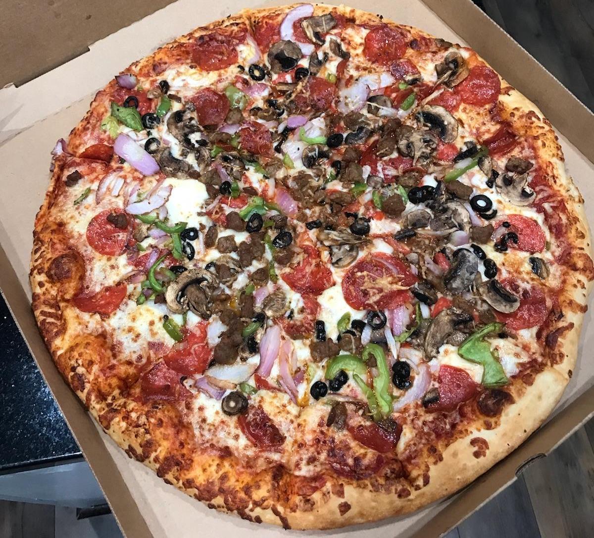 A large pizza in an open box, topped with pepperoni, mushrooms, green peppers, onions, black olives, and sausage, divided into multiple slices.