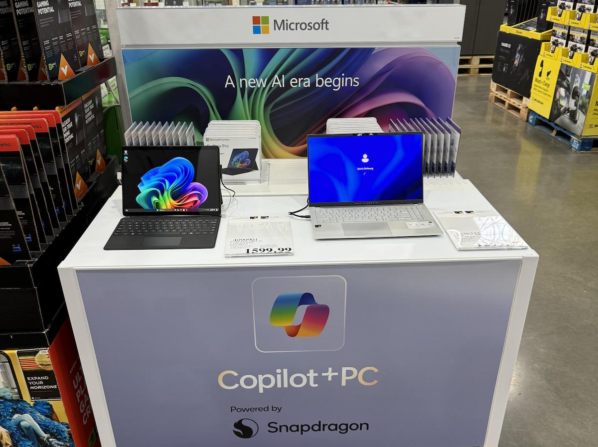 A retail display for Microsoft Copilot+ PCs powered by Snapdragon, featuring two open laptops and a colorful sign reading “A new AI era begins.” The display is set up in a store with shelves in the background.