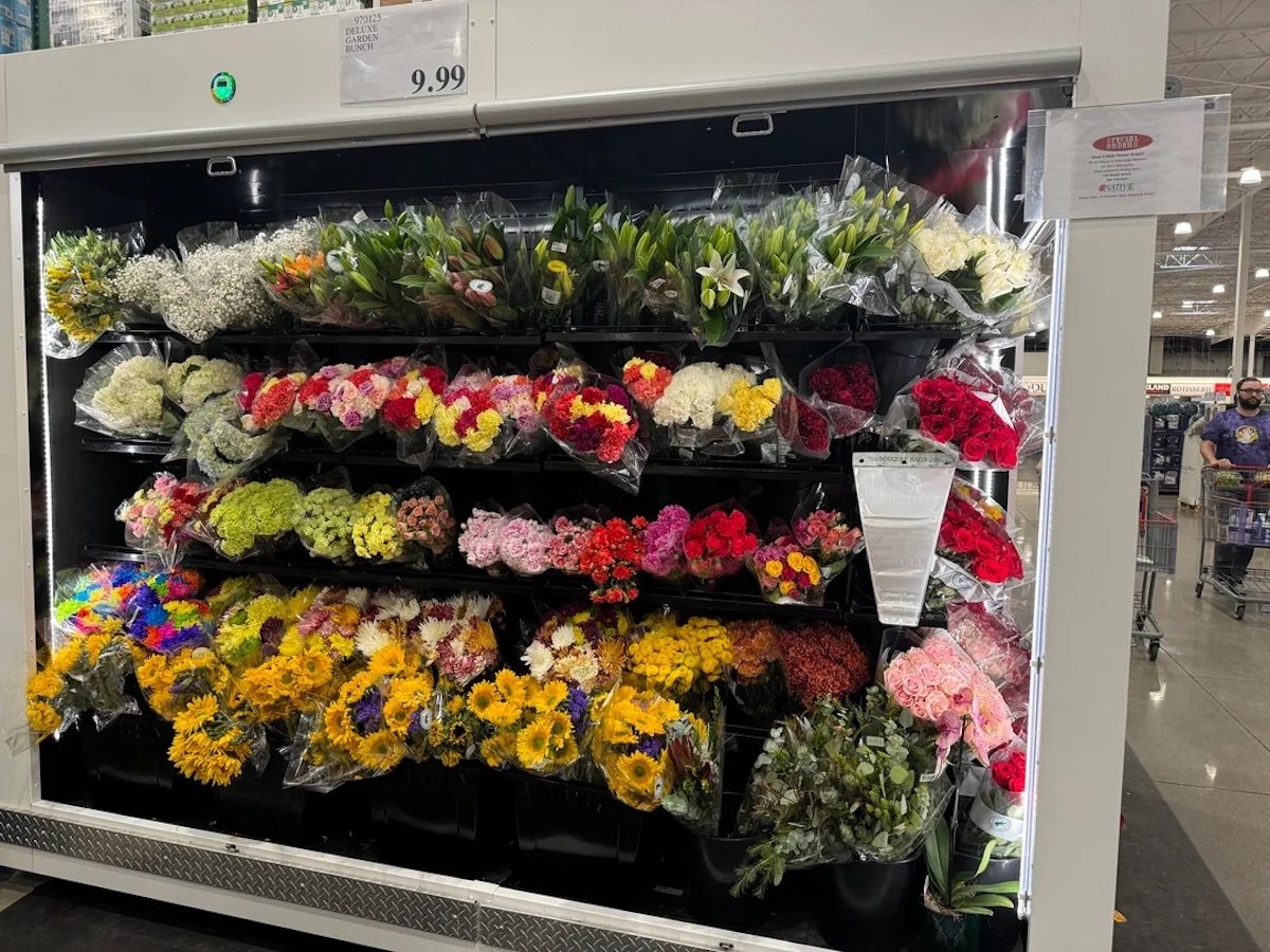 A refrigerated display case in a store holds multiple shelves of assorted fresh flower bouquets, including roses, lilies, sunflowers, and mixed blooms in various colors. A sign above shows a price of $9.99.