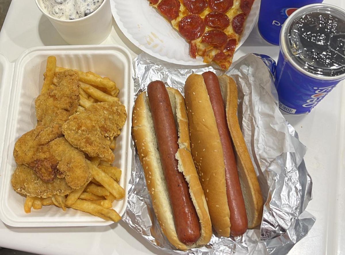 A table with fried chicken and fries in a container, two hot dogs on foil, a slice of pepperoni pizza on a paper plate, a cup of soda, and a cup of cookies and cream ice cream.