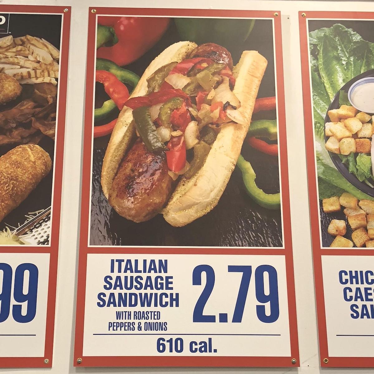 A poster displays an Italian sausage sandwich topped with roasted peppers and onions in a bun, priced at $2.79 and labeled as 610 calories. The sandwich is set against a background of sliced peppers.