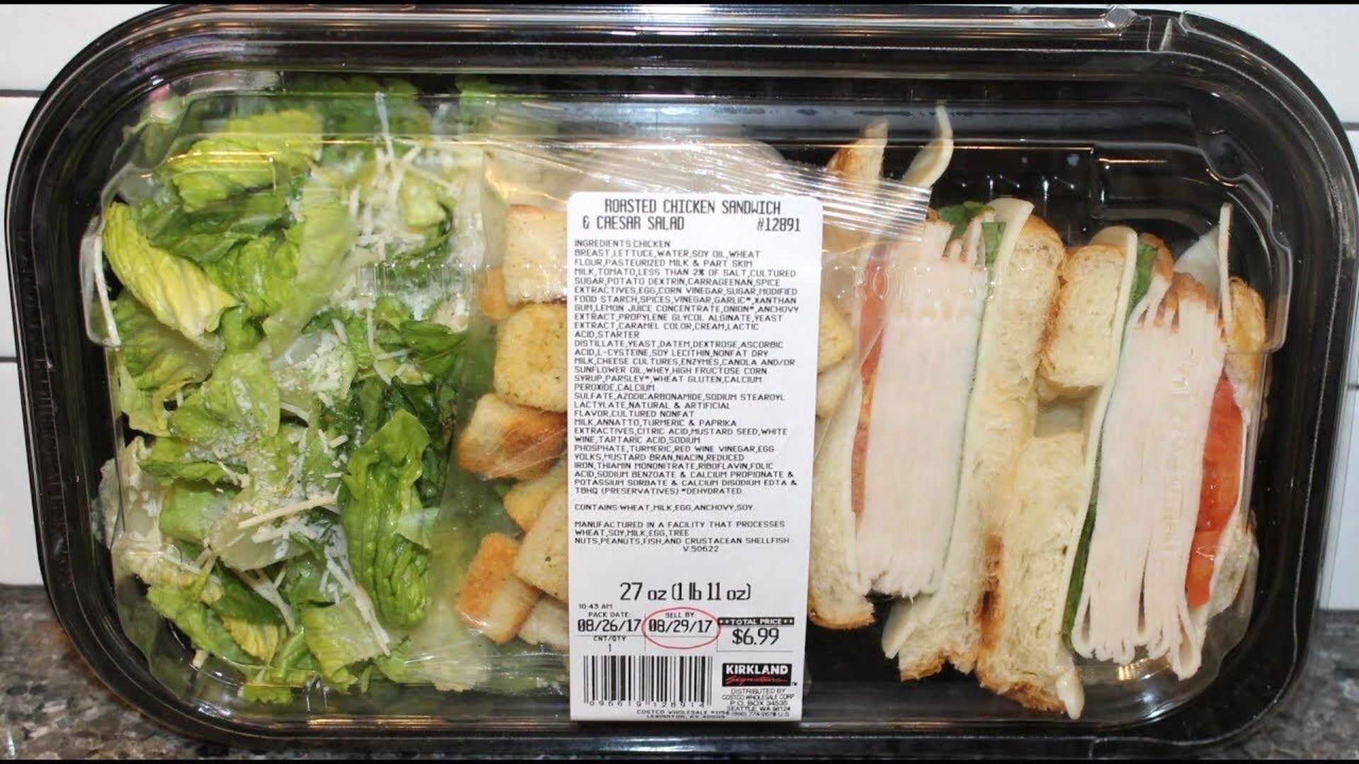 A plastic container with a Caesar salad on the left, topped with croutons and grated cheese, and sliced turkey sandwiches with tomato, lettuce, and cheese on the right. A label with nutrition facts is in the center.