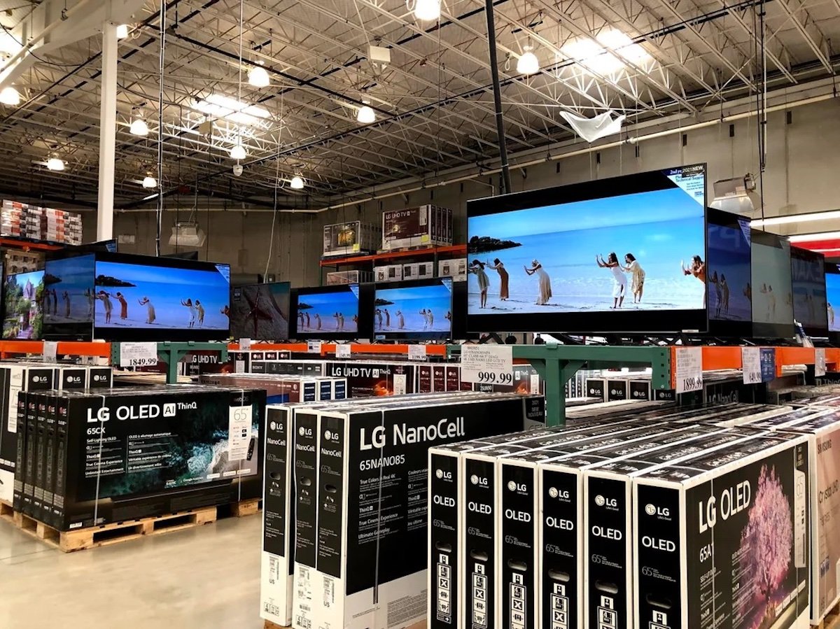 Large warehouse store electronics section with several LG TVs on display, all showing the same beach scene from a movie. Boxes of LG OLED and NanoCell TVs are stacked below the displays. Bright overhead lighting above.