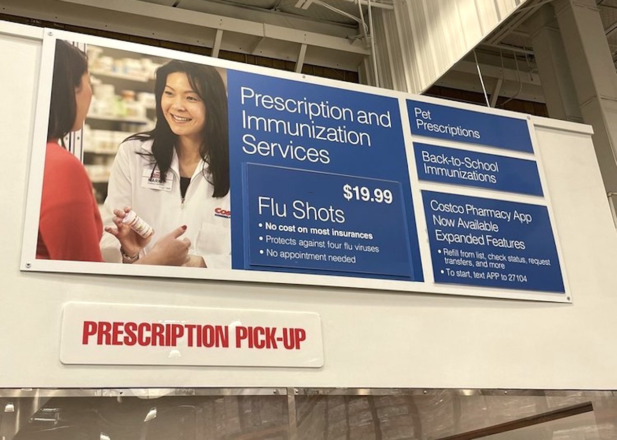 A sign at a pharmacy advertises prescription and immunization services, including $19.99 flu shots, pet prescriptions, back-to-school immunizations, and pharmacy app features. Below is a "Prescription Pick-Up" sign.