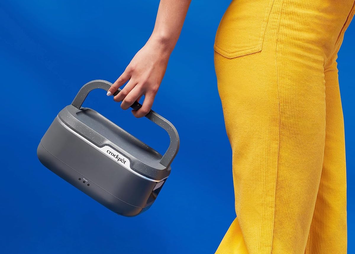 A person dressed in bright yellow pants holds a portable gray speaker or device by its handle against a solid blue background.
