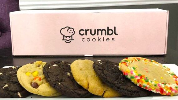 A row of assorted cookies sits on a plate in front of a pink Crumbl Cookies box. The cookies vary in flavors, including chocolate, peanut butter, and one with colorful sprinkles.
