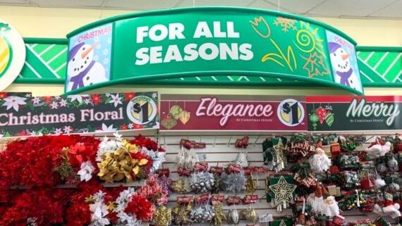A store display labeled “FOR ALL SEASONS” features Christmas floral decorations, ornaments, and festive items under signs reading “Elegance” and “Merry,” with various red, gold, and green holiday decorations.