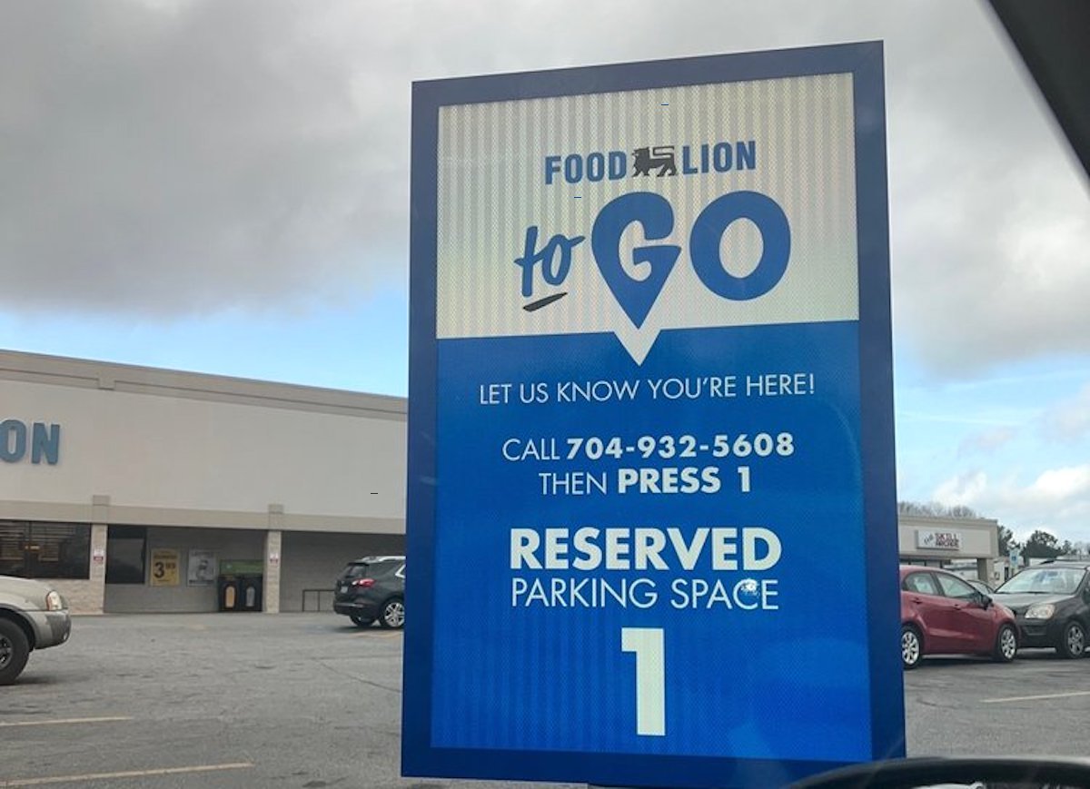 A blue sign in a grocery store parking lot reads: “Food Lion To Go. Let us know you’re here! Call 704-932-5608 then press 1. Reserved Parking Space 1.” The store building is visible in the background.